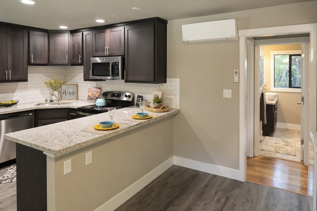 Kitchen with stainless appliances, stone counters, tile backsplash, wood look flooring, brown cabinets and view into bathroom