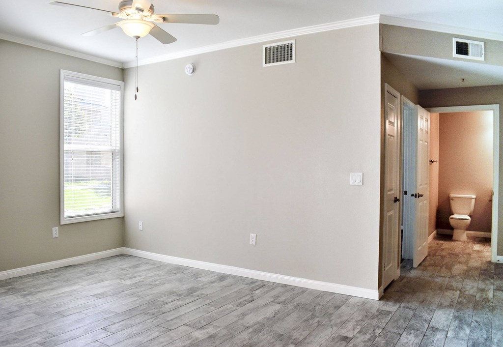 View from living room with well lit window and ceiling fan towards hall with bathroom, has wood look flooring