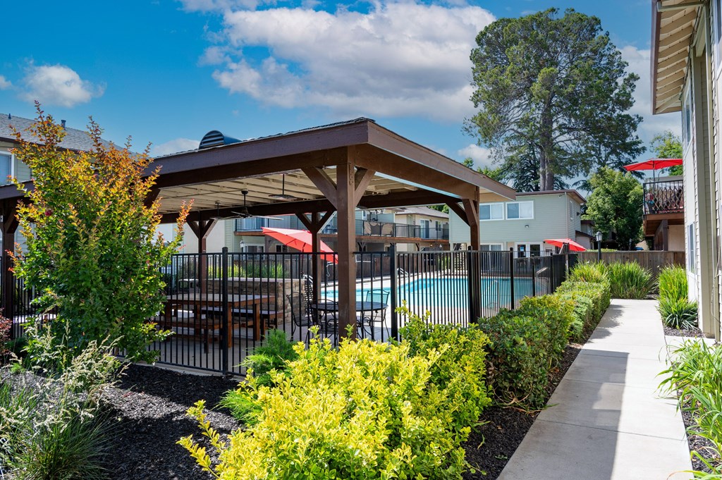 View into gated community pool with covered area that has a picnic table and seating areas, surrounded by lush landscaping