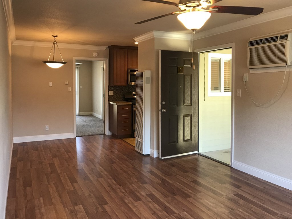 View from living area into kitchen and bedroom. Living area has wood look flooring and ceiling fan, kitchen has tile flooring, and bedroom has carpet flooring