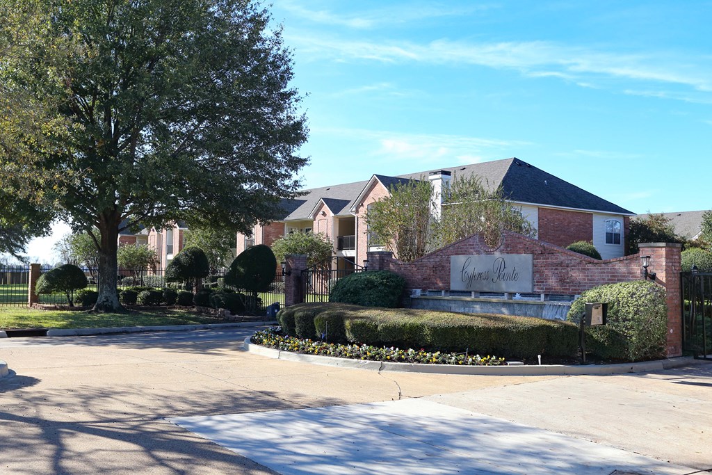 View of front entry gate with lush shrubbery, waterfall below logo on gate, view of landscaping and exterior of buildings.