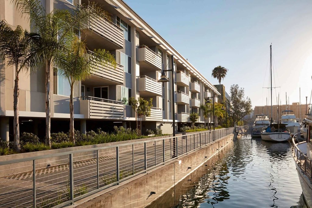 A row of modern buildings line a canal with boats docked at the marina.