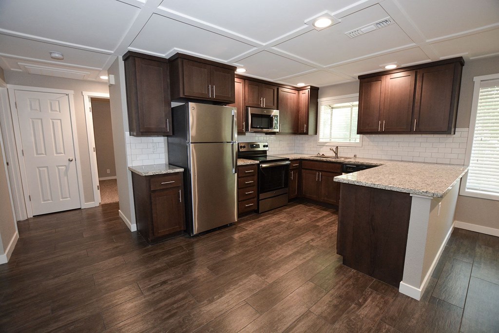 View of open kitchen with stone counters, tile backsplash, stainless appliances, brown cabinets, recessed lighting, and wood look flooring