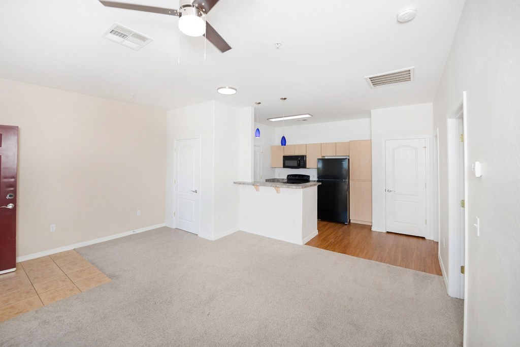 A spacious living room with a ceiling fan and a kitchenette in the background.