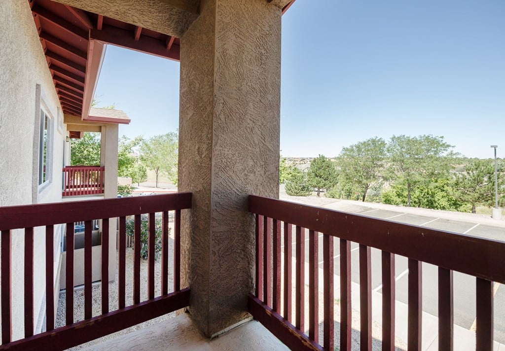 A balcony with a red railing and a view of a parking lot and trees.