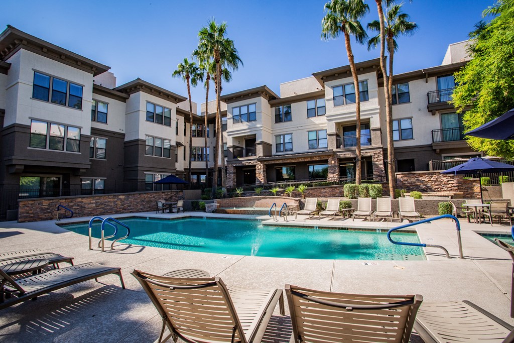A swimming pool surrounded by lounge chairs and palm trees in front of a multi-story apartment building.