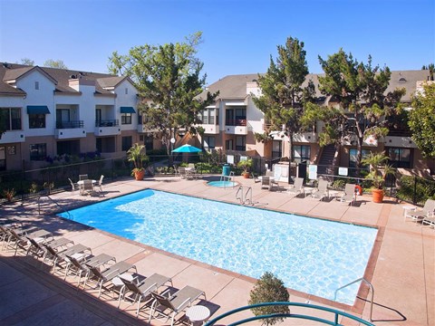 Ventura, CA, Apartments - Cypress Point - Sparkling Pool with Spacious Sundeck, Hot Tub, Lounge Seats, Umbrella Tables and Chairs, Surrounding Trees, and Apartment Buildings in the Background.