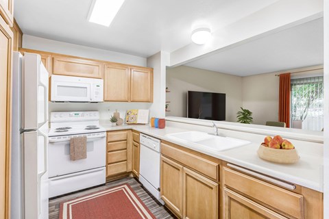 Goleta, CA Apartments Near Costco - Encina Meadows - Kitchen with Plank Flooring, Rug, White Appliances, Wood-Style Cabinetry, Breakfast Bar, and View of Living Room.