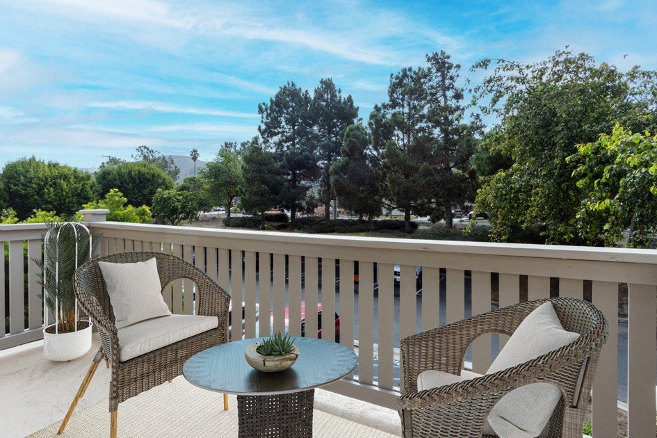 Two-Bedroom Apartments in Lompoc, CA - Oceanwood - Private Balcony with Patio Chairs, Coffee Table, Potted Plant, and View of Parking Lot with Lush Trees in the Background.