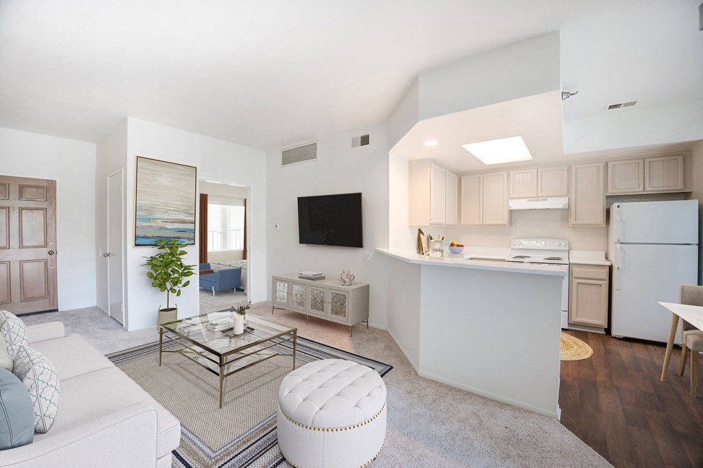 Pet-Friendly Apartments in Santa Barbara, CA - Rancho Franciscan - Living Room with Carpet Flooring, Couch, Ottoman Stool, Glass Coffee Table, Storage Cabinet, Wall-Mounted Television, and View of Kitchen and Bedroom.