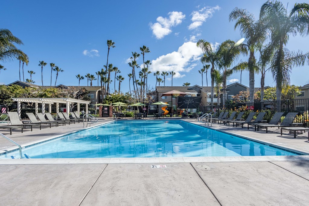 Apartments for Rent in Goleta, CA - Sumida Gardens - Pool with Spacious Sundeck, Lounge Seats, Umbrella Tables with Chairs, Pergola, and Palm Trees.