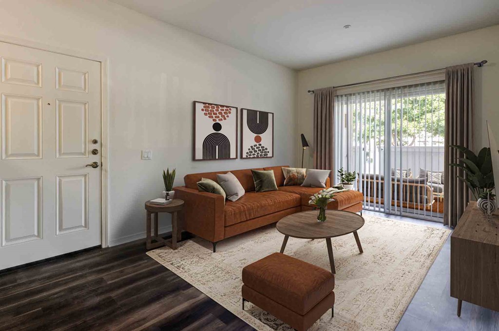 Goleta CA Apartments - Sumida Gardens - Furnished Living Room with Wood-Style Flooring, Area Rug, Burnt Orange Sofa, Ottoman, Wooden Round Coffee Table, Side Table with Plant, Wooden Monitor, Standing Lamp, Framed Art, and Glass Sliding Door with Access to Balcony