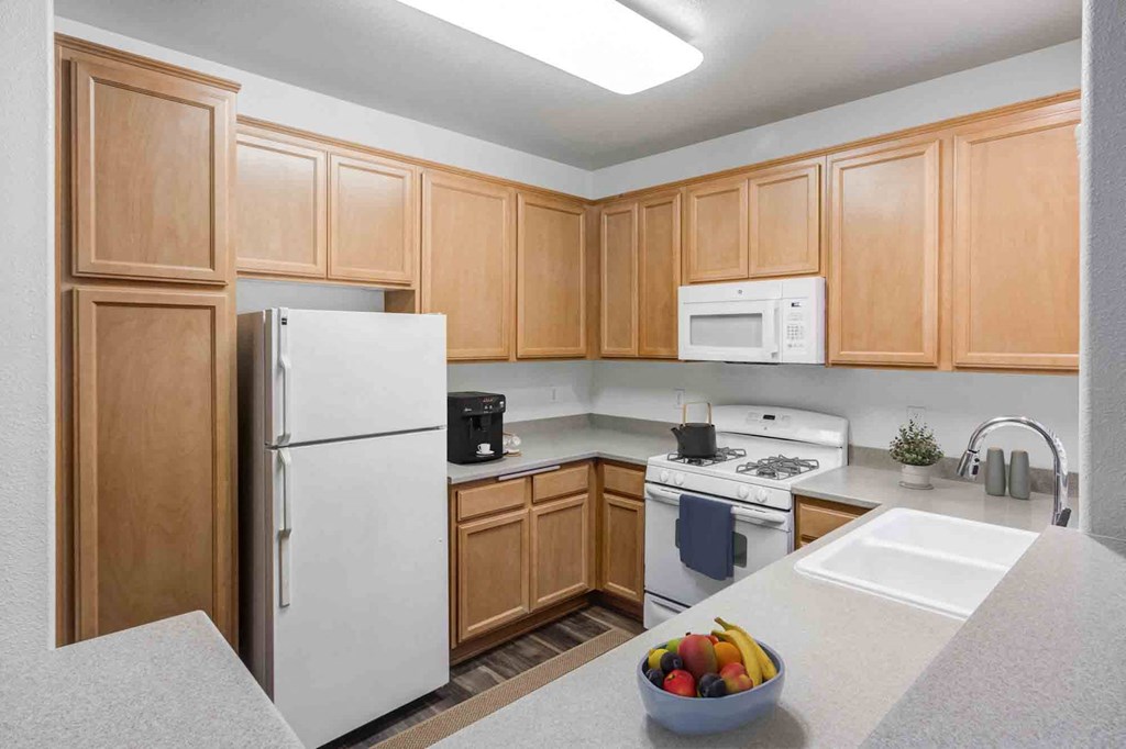 Apartments for Rent in Goleta CA - Sumida Gardens - Kitchen with Wood-Style Floor, Area Rug, White Appliances, Wood Cabinetry, Grey Countertops, Overhead Lighting, and Breakfast Bar Area