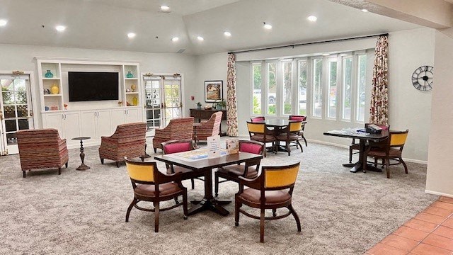 Santa Barbara, CA, Apartments Near Target - Rancho Franciscan - Community Clubhouse With Tables and Chairs, Carpeting, Mounted TV, Bookshelf, and Large Windows