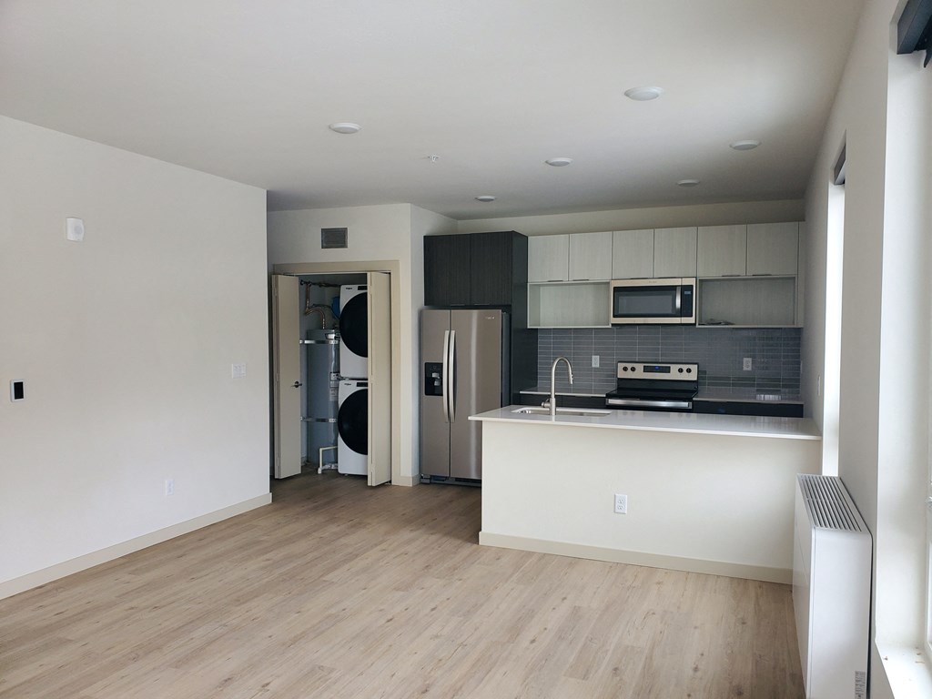 an empty living room with a kitchen with a refrigerator and a sink