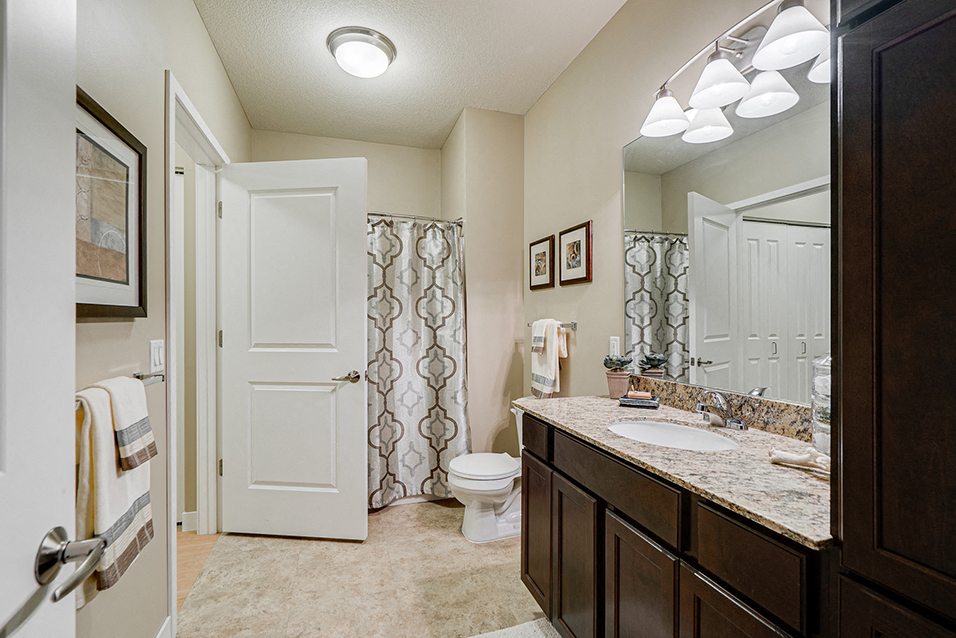 Bathroom with bright lights, brown cabinets, and light granite countertops