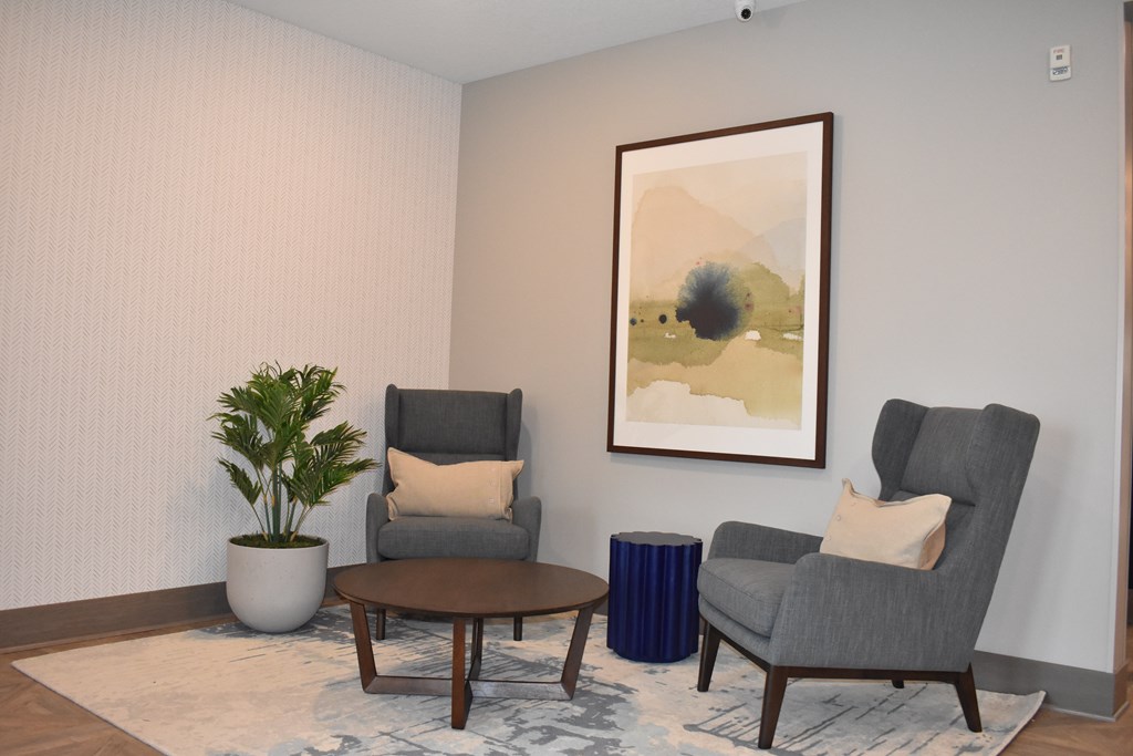 A living room with two grey chairs, a table, a potted plant and a painting on the wall.