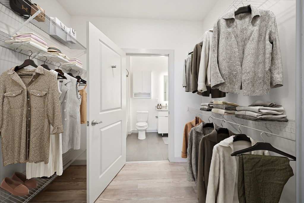 A walk in closet with clothes hanging and a toilet in the background.