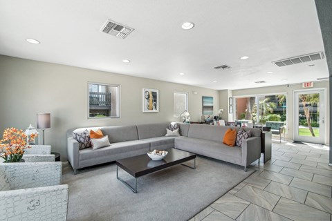 A grey couch with orange pillows sits in a living room with a glass coffee table in front of it.