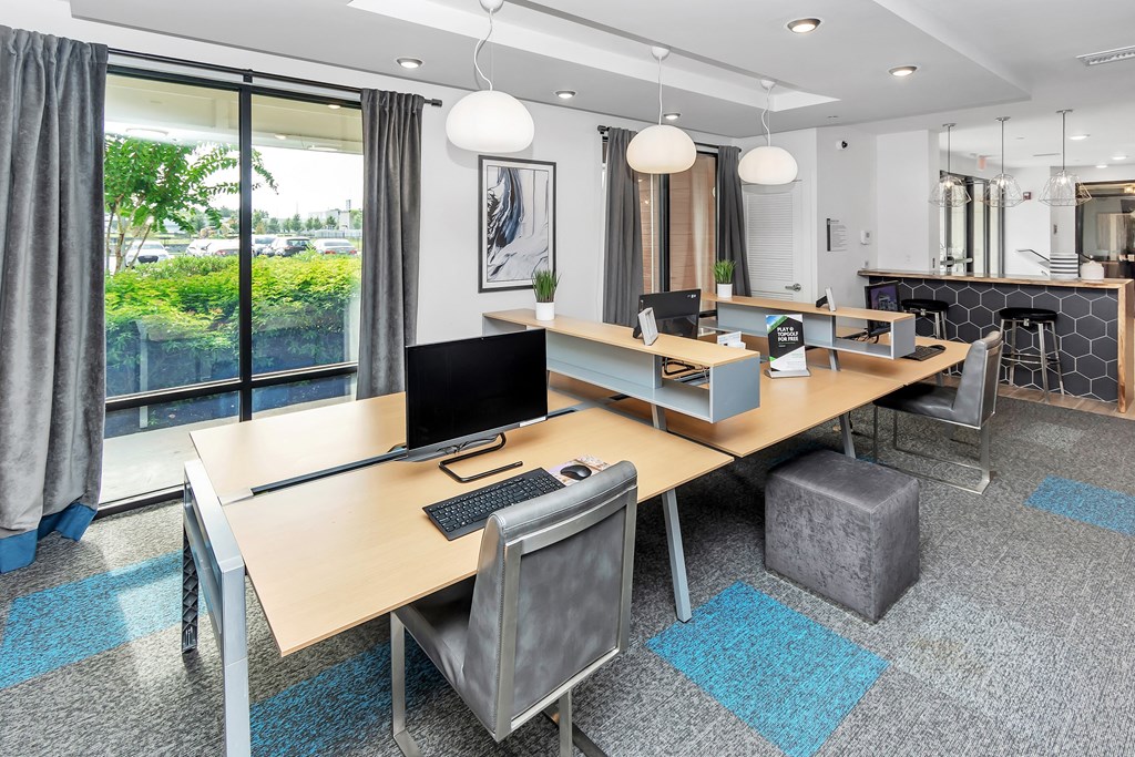 a room with desks and chairs with a large window and a kitchen in the background