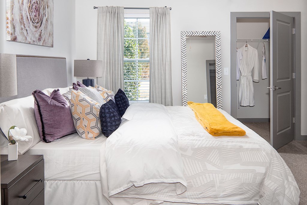 A bed with a white comforter and a yellow pillow is in the foreground of a room with a closet and a mirror.