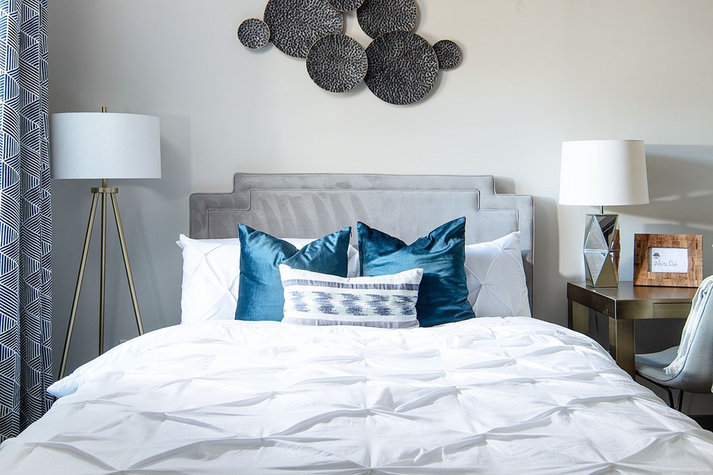 A bed with white sheets and blue pillows is in the foreground with a lamp and a picture frame on a side table.