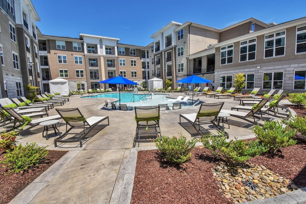 A pool surrounded by chairs and umbrellas in front of apartment buildings.