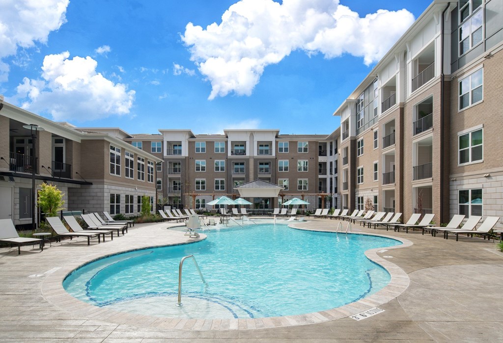 A large swimming pool surrounded by lounge chairs and umbrellas in front of apartment buildings.