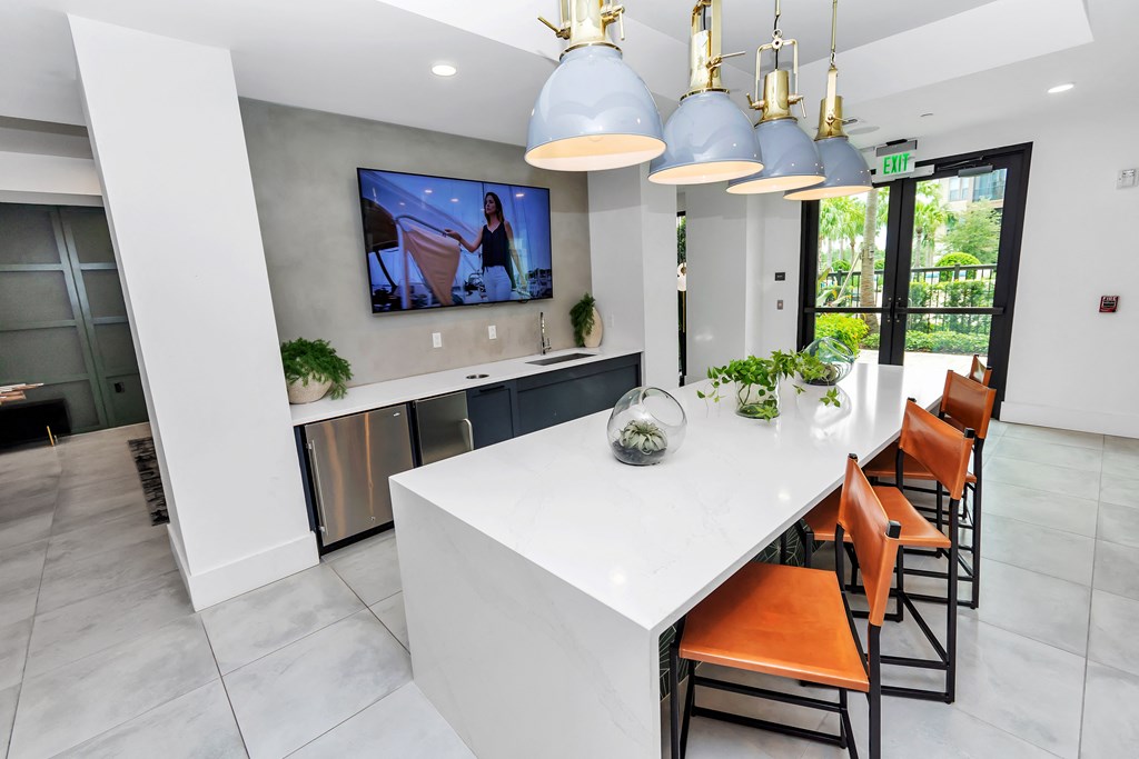 a kitchen with a large white island with orange chairs and a large tv on the wall