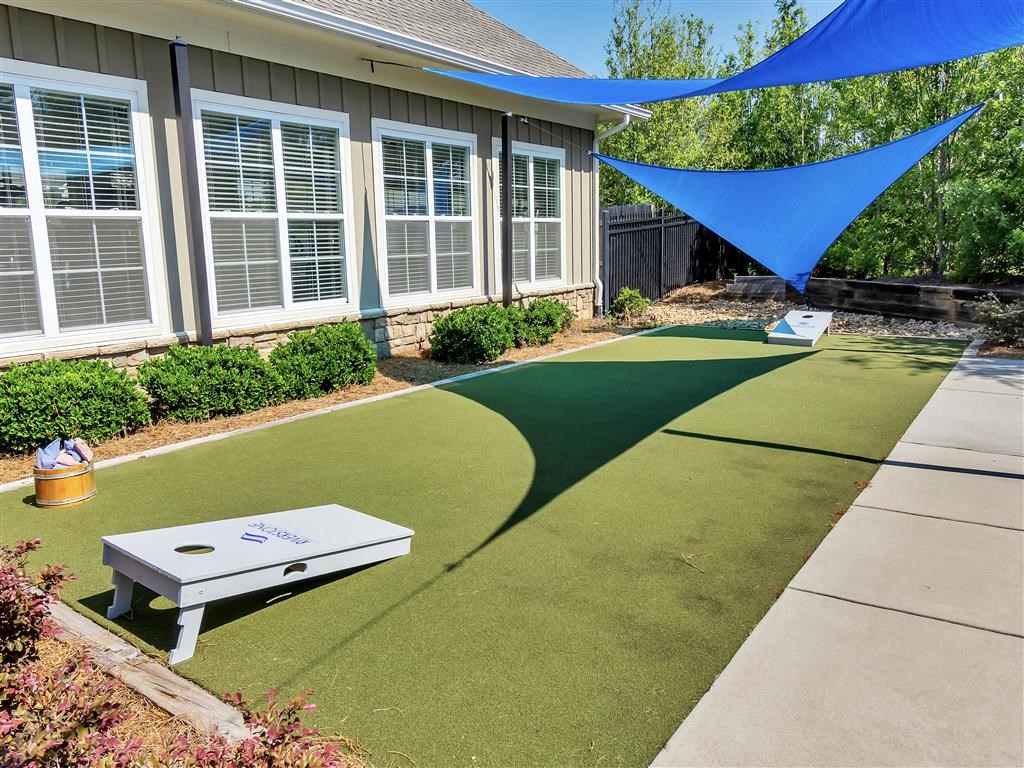 Outdoor grassy area with a cornhole game under triangular shade sails hanging above near a building