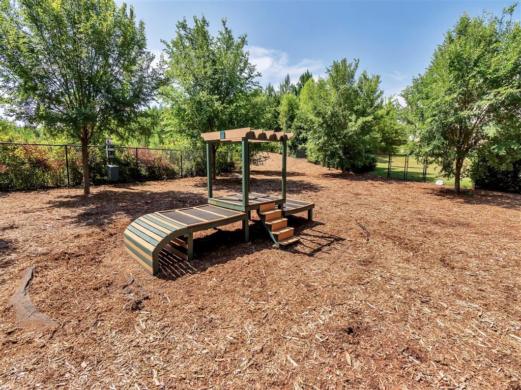 Gated dog park with mulch, trees, and a piece of agility equipment with steps and a raised surface