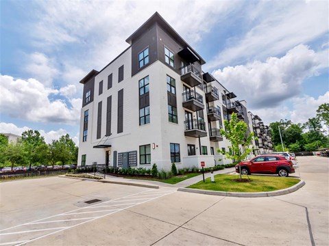 A modern four-story apartment building with balconies near a parking lot with cars and trees