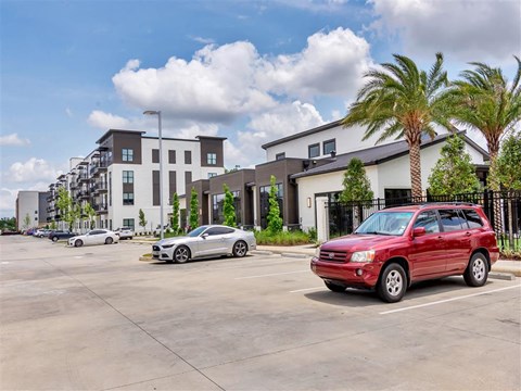 Parking area with cars near a landscaped community clubhouse and a row of apartment buildings