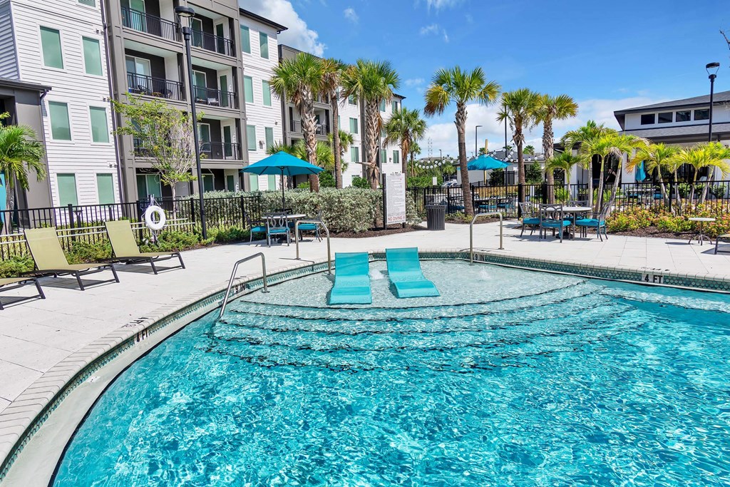 a swimming pool with blue lounge chairs and umbrellas in front of an apartment building