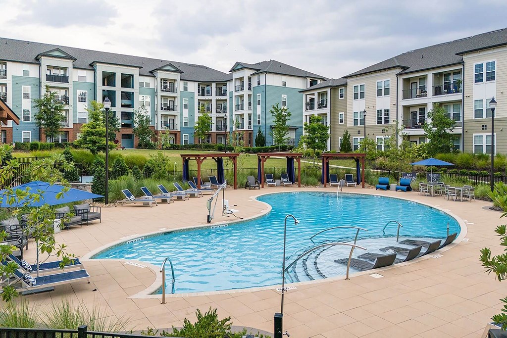 A large swimming pool surrounded by lounge chairs and umbrellas in front of apartment buildings.