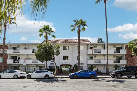 Cars parked in front of a white apartment building with palm trees.