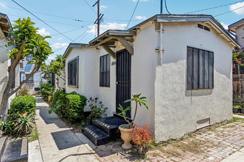 A small white house with a black door and windows surrounded by plants and a brick walkway.