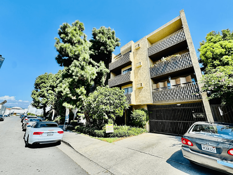A street view of a residential area with cars parked on the side of the road and a building with balconies in the background.