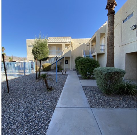 A concrete pathway leads to a building with a blue fence and palm trees.