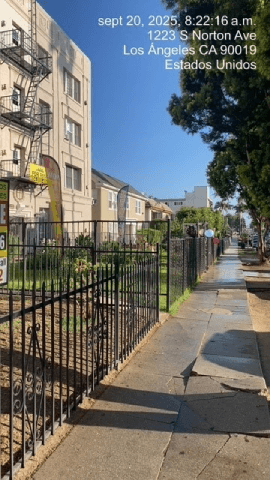 A black metal fence runs along a sidewalk in front of apartment buildings.