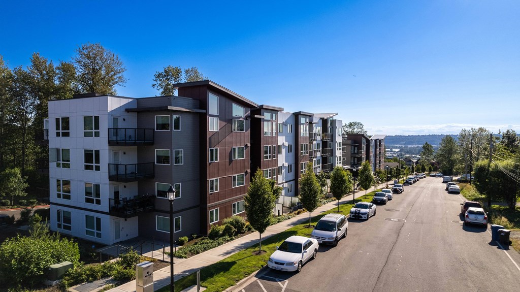 Drone shot of the apartment building showcasing the exterior of the building and the neighborhood. Cars parked along the street  in front of the building and clear blue skies outside.