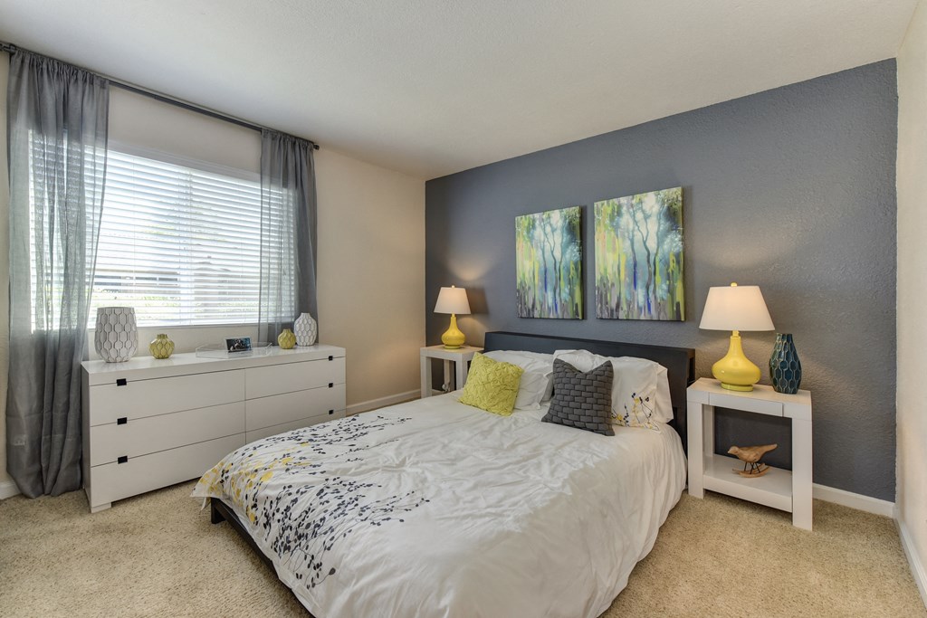 Bedroom with Large Window, White Comforter on Mattress, Lamps and Carpet at Monte Bello Apartments, Sacramento