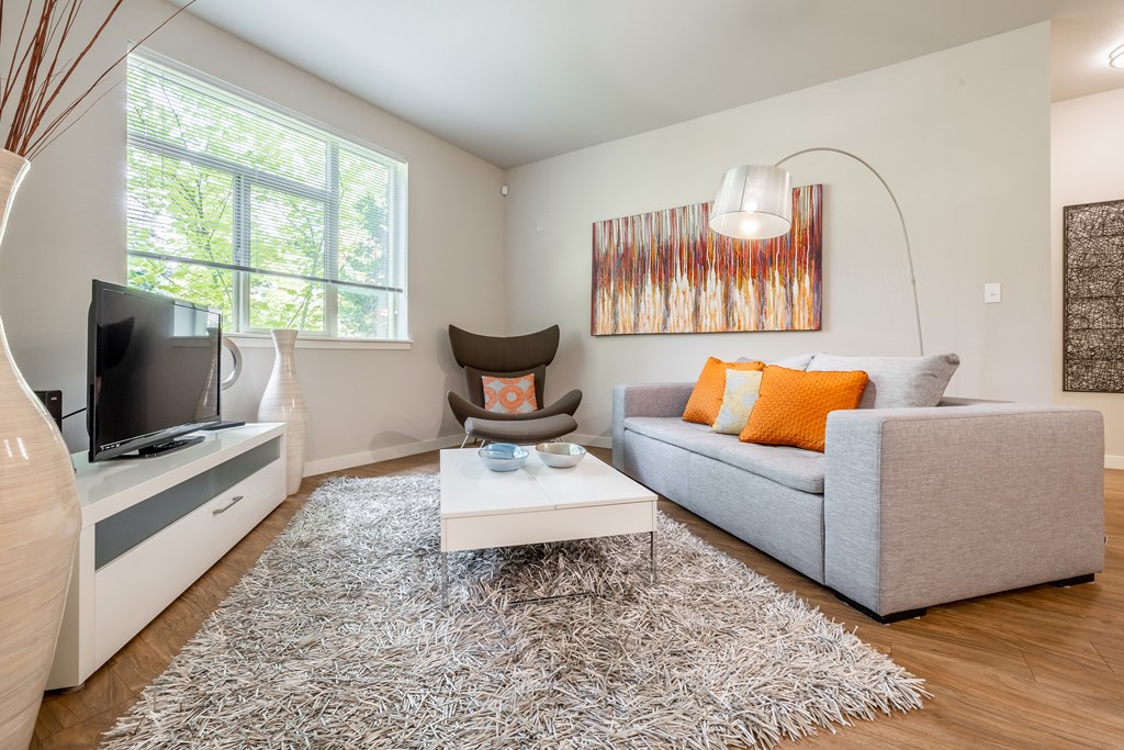 Model home living room with light grey couch, lamp that hangs over the couch, windows and chair in the corner.  Flooring is wood with an area rug in the center