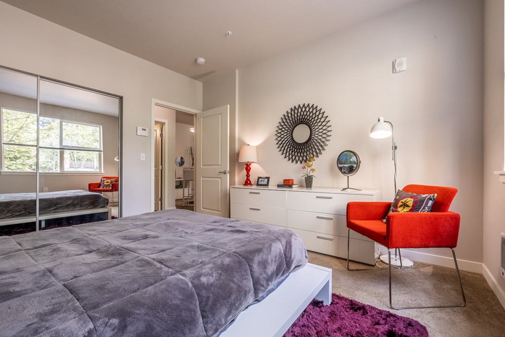 Bedroom with mirrored closet doors, carpet throughout with a burgundy accent rug and long white dresser on the wall opposite of bed.