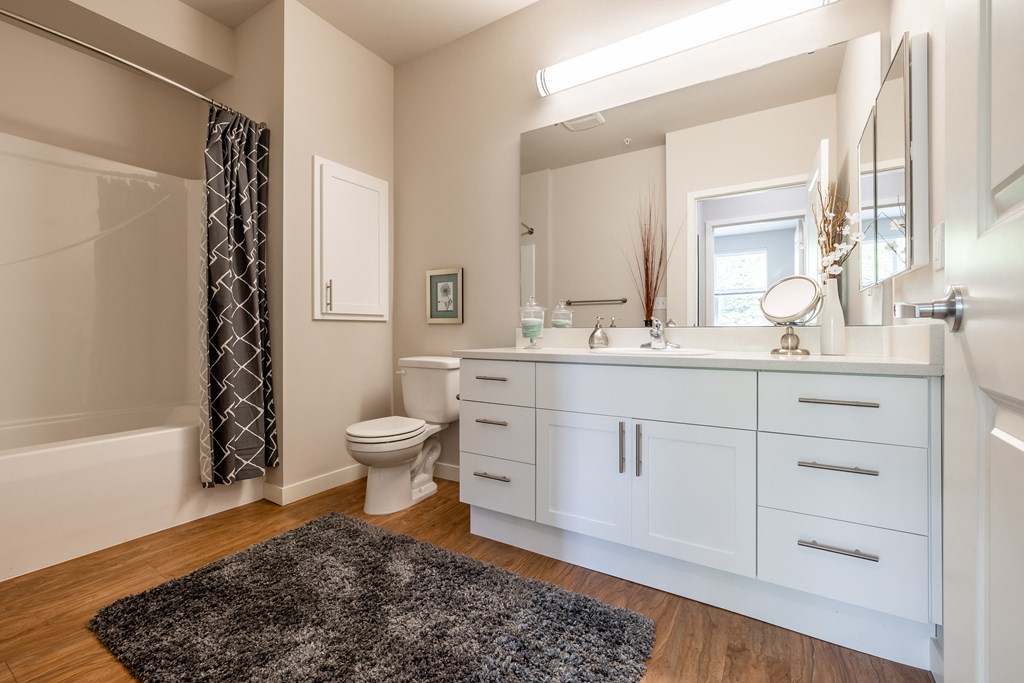 Model Home furnished bathroom with gray rug, white cabinets, gray shower curtain hanging at the tub/shower enclosure.