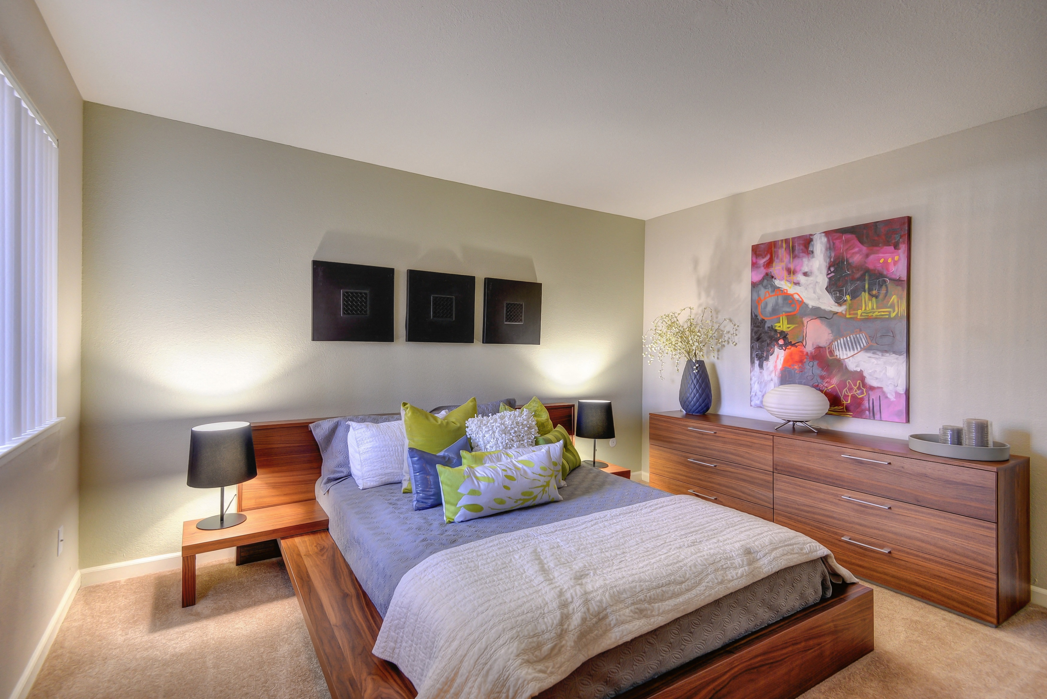 Large Bedroom with Full Sized Bed on Elevated Wood Frame, Carpet, Wood Dressers, Small Black Lamps on each side of Bed, Abstract Painting on Wall at Addison Ranch Apartments, Petaluma, California
