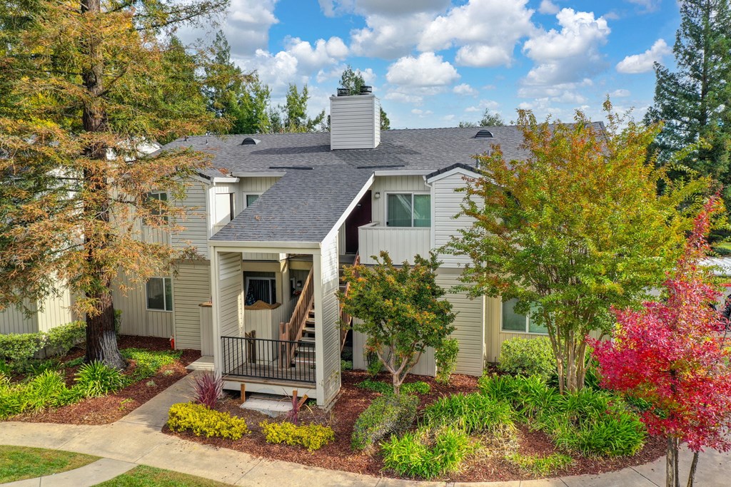 Property apartment building that is two stories.  There are mature trees and shrubs outside of the building. One tree is red with fall colors. at Monte Bello Apartments, California