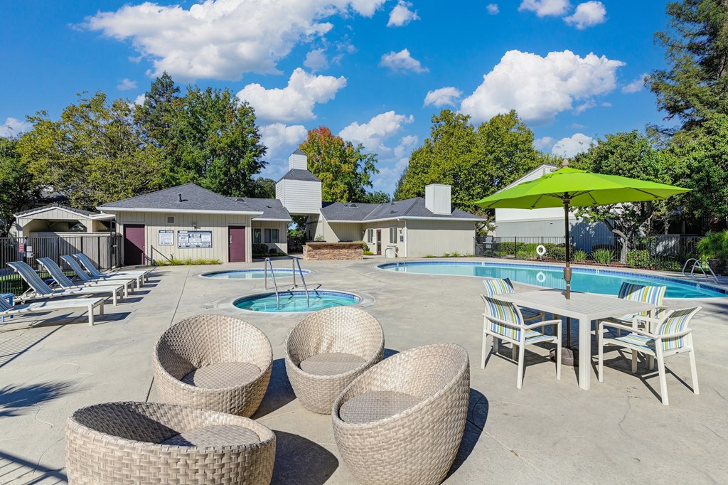 Swimming pool deck with round wicker-like lounge chairs.  There is a square table, chairs and line green umbrella for additional seating around the swimming pool at Monte Bello Apartments