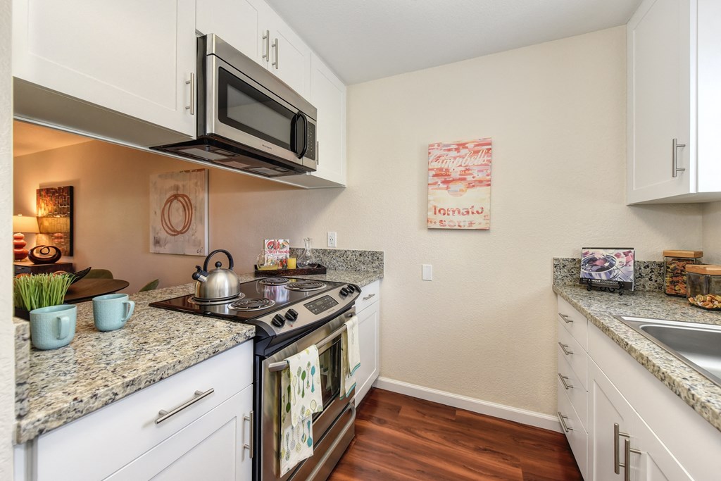 Model home kitchen with granite counter tops, white cabinets and wood flooring.  Kitchen comes with refrigerator, microwave, stove and dishwasher. at Monte Bello Apartments, Sacramento, California