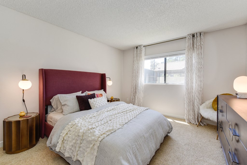 Model home bedroom with Queen size bed with burgundy colored headboard, wooden bedside table with lamp, carpet through out and a large window